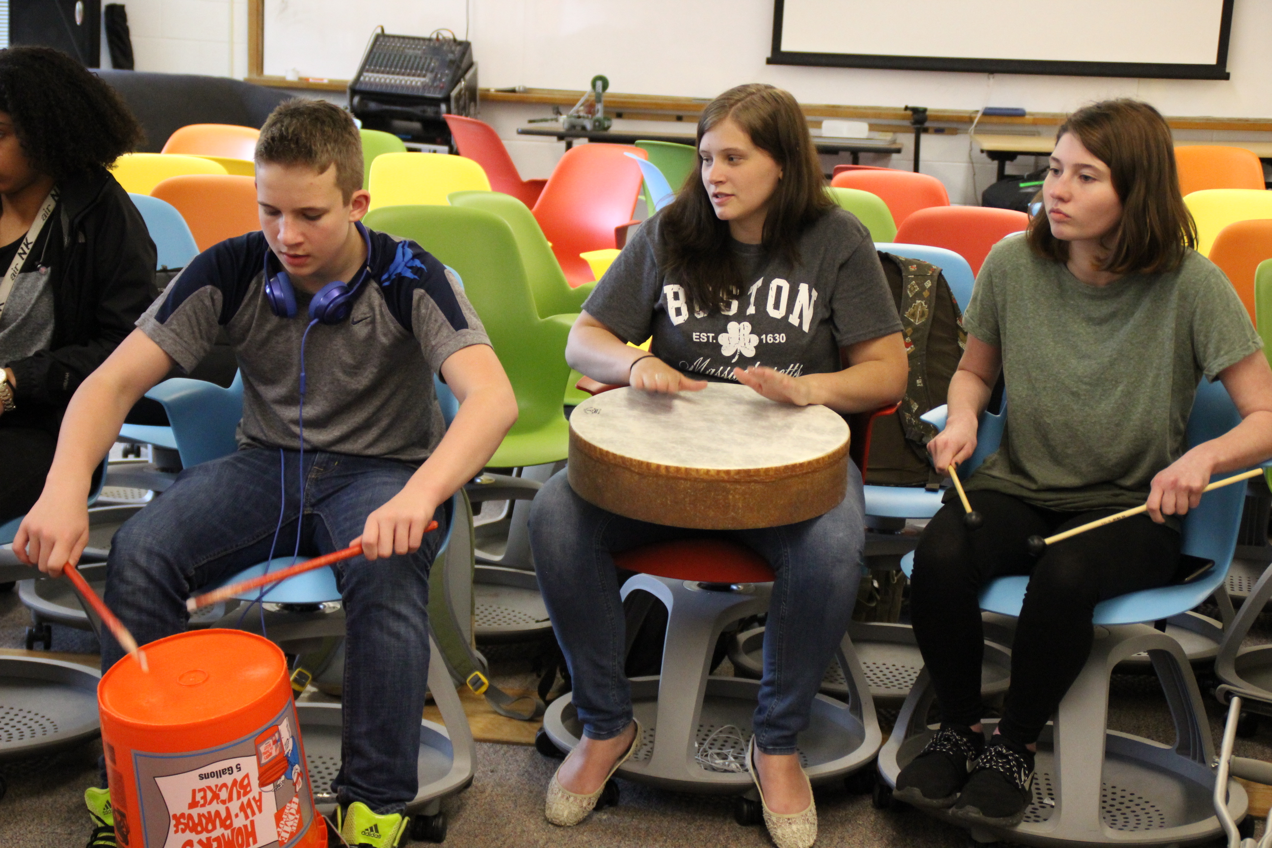 A students uses drumstick on a bucket, another uses her hands on a lap drum and a third students uses drumsticks on her legs