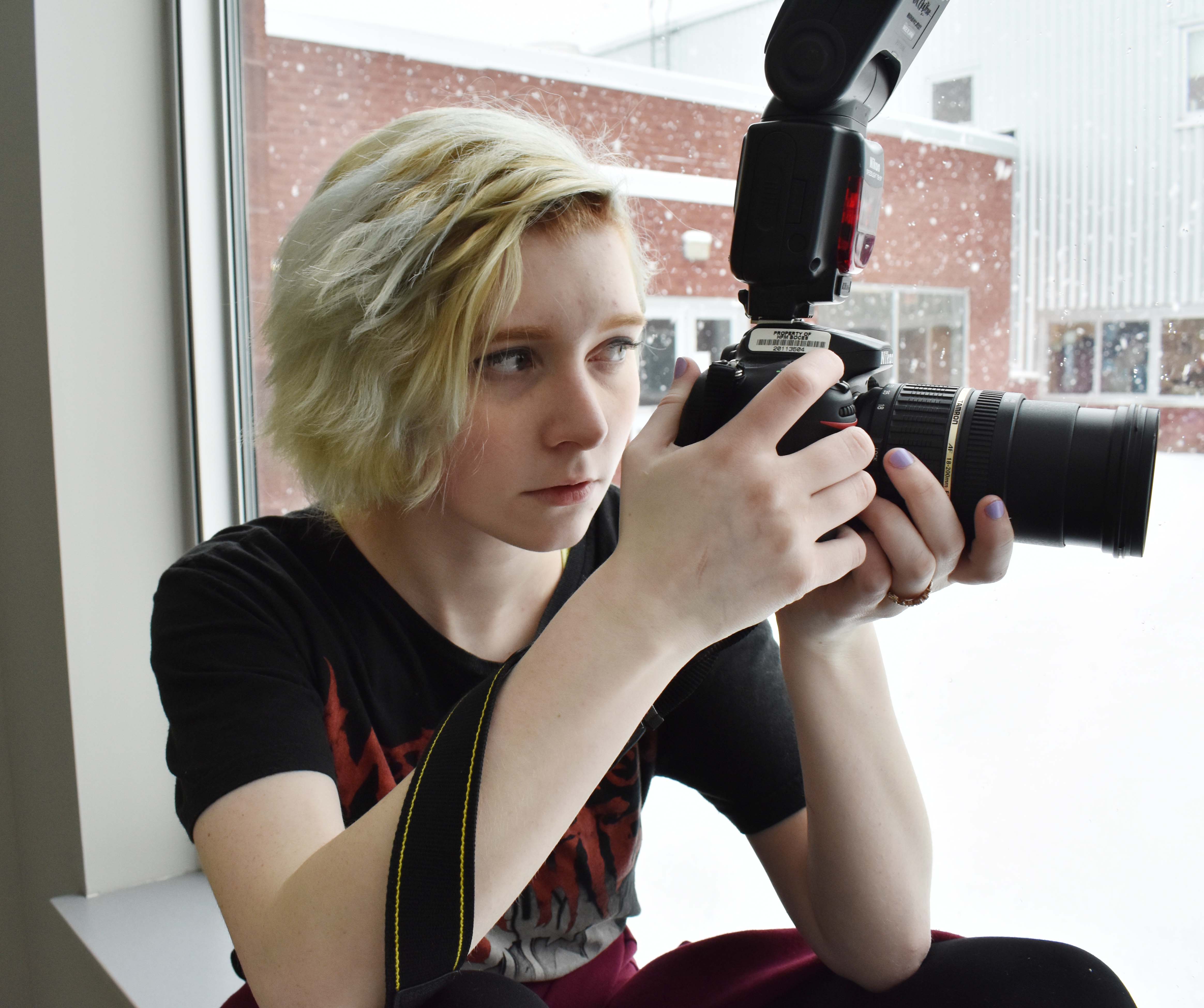 A student with a camera sits near a window preparing to take a picture.