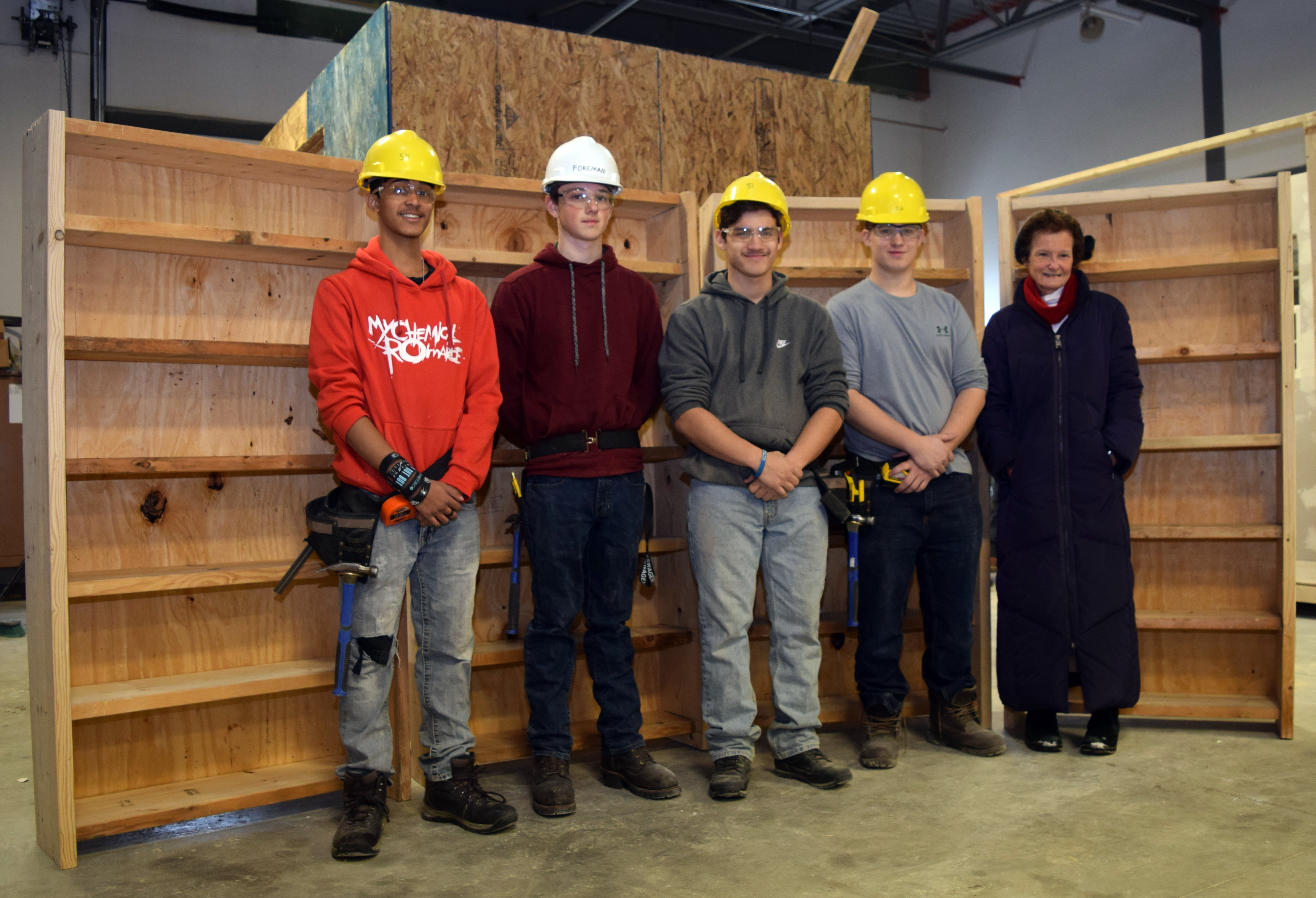Four students wearing hard hats and an adult stand in front of four six-foot tall bookcases.