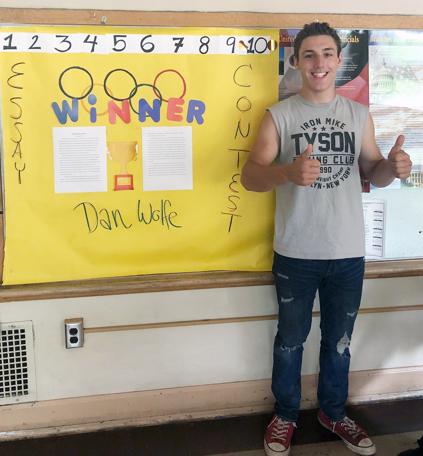 Daniel gives two thumbs up while standing next to a bulletin board announcing his essay contest win.