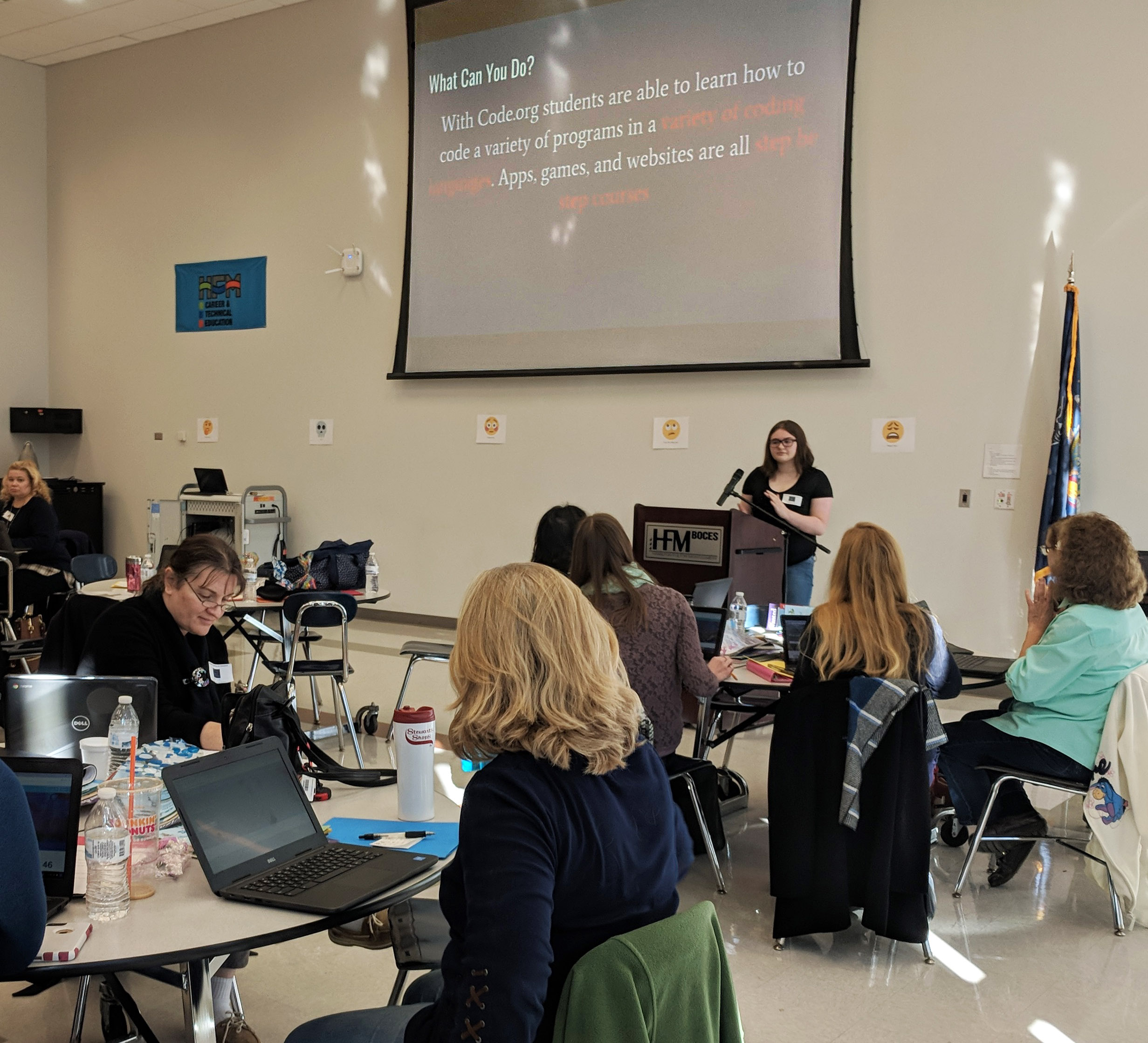 A student stands at a podium with a Powerpoint in the background as she presents to a crowd of people seated at tables. 