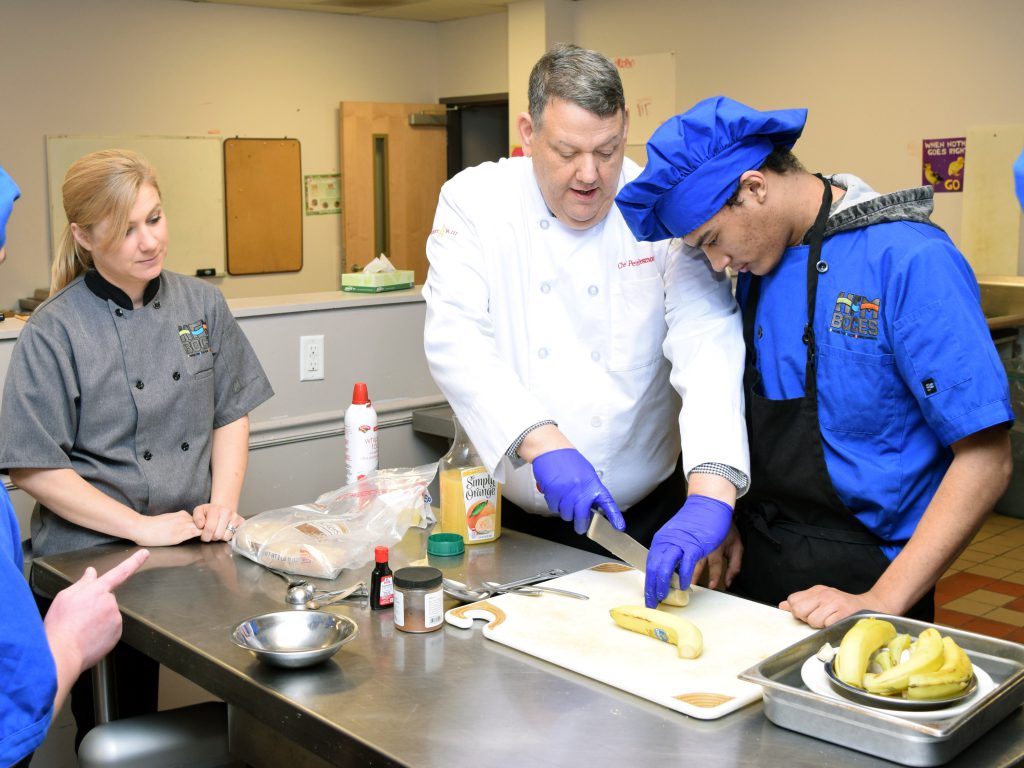 An instructor in a while culinary coat sits cuts a banana as a student and another instructor look on.