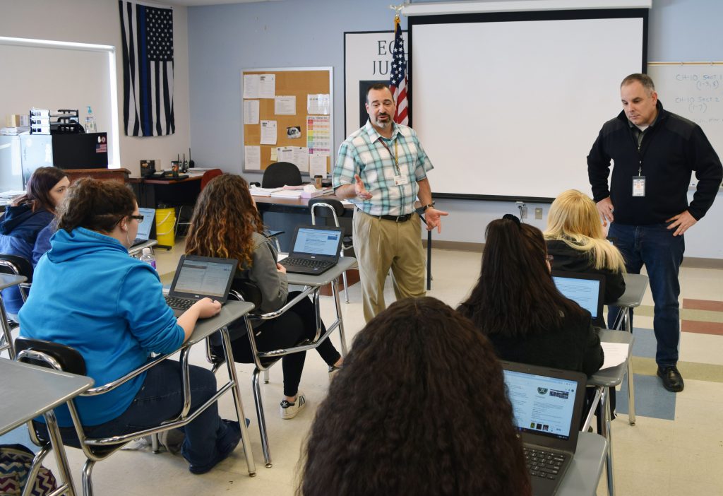 Two instuctors stand in front of a classroom talking with students sitting at desks.