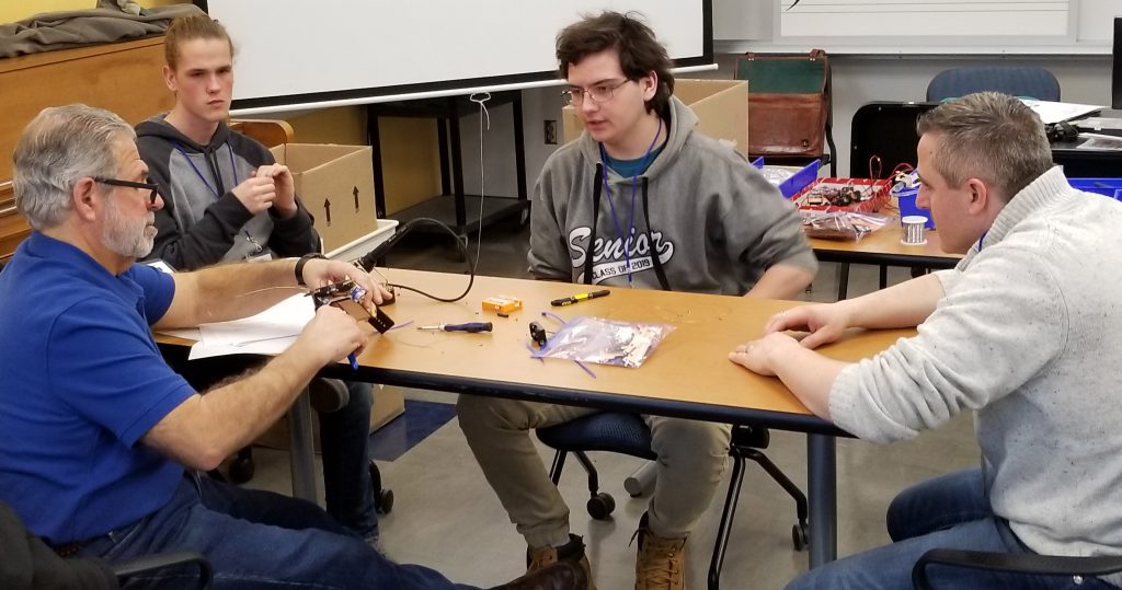 four people sit around a table assembling a small robot.