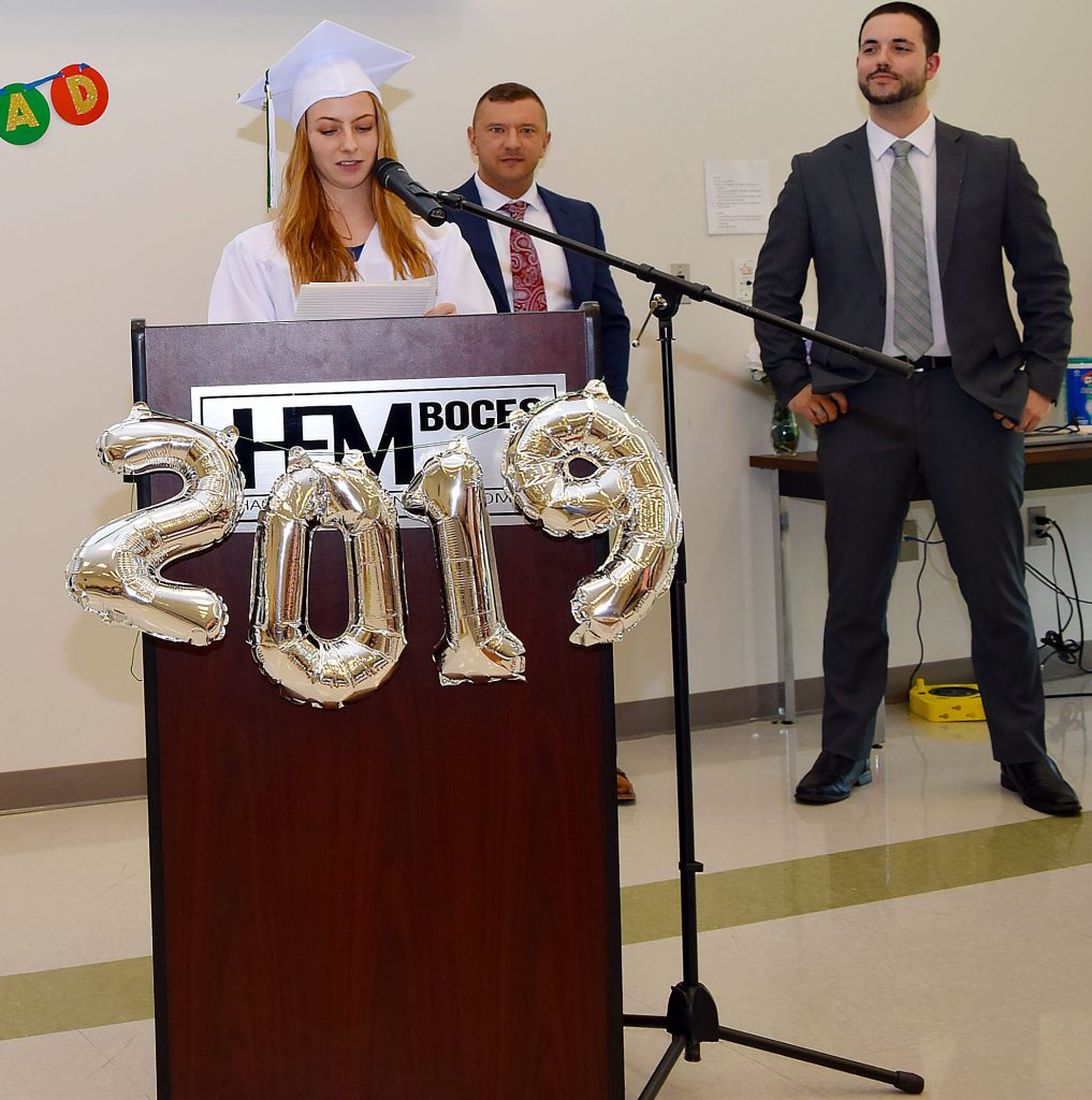 A student in a cap and gown addresses an audience from a podium. Two teachers are in the background.