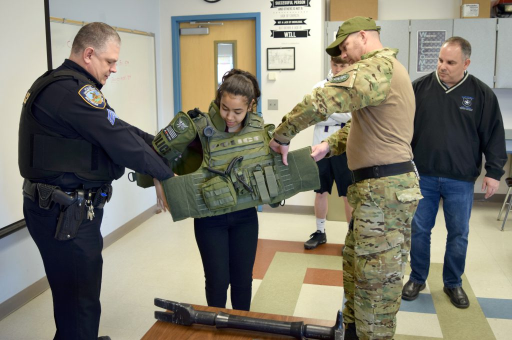 Two police officers help a student into a bullet proof vest as the instructor looks on.