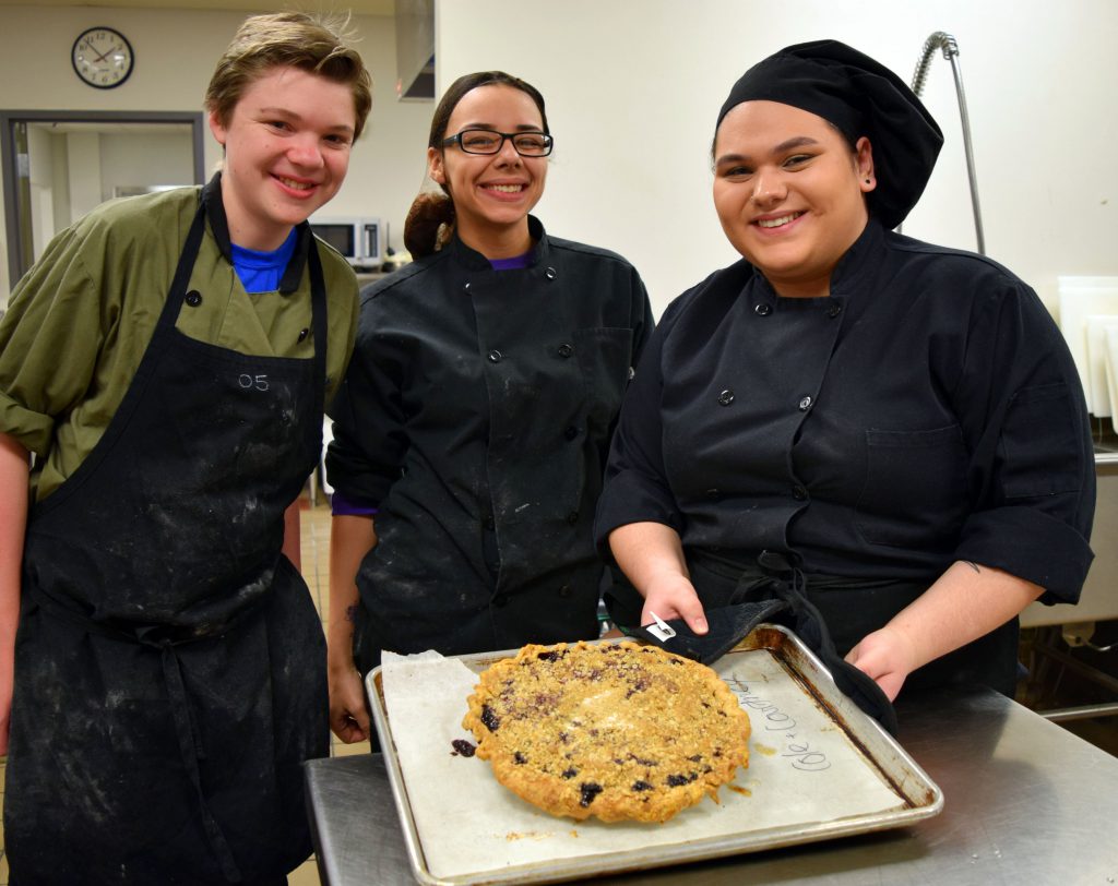 Three students in culinary uniforms display a pie on a baking sheet