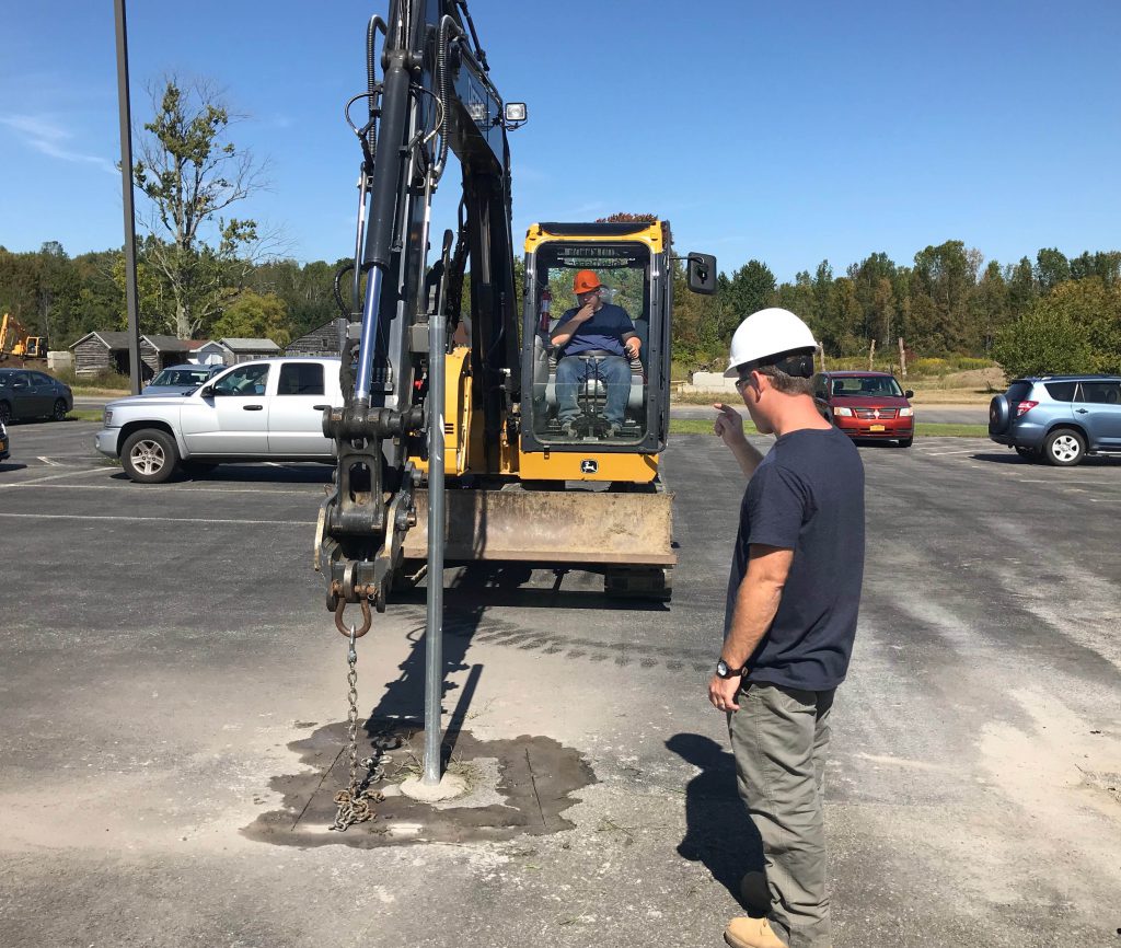 A student uses an excavator and chain to pull and metal fence post out of the ground.
