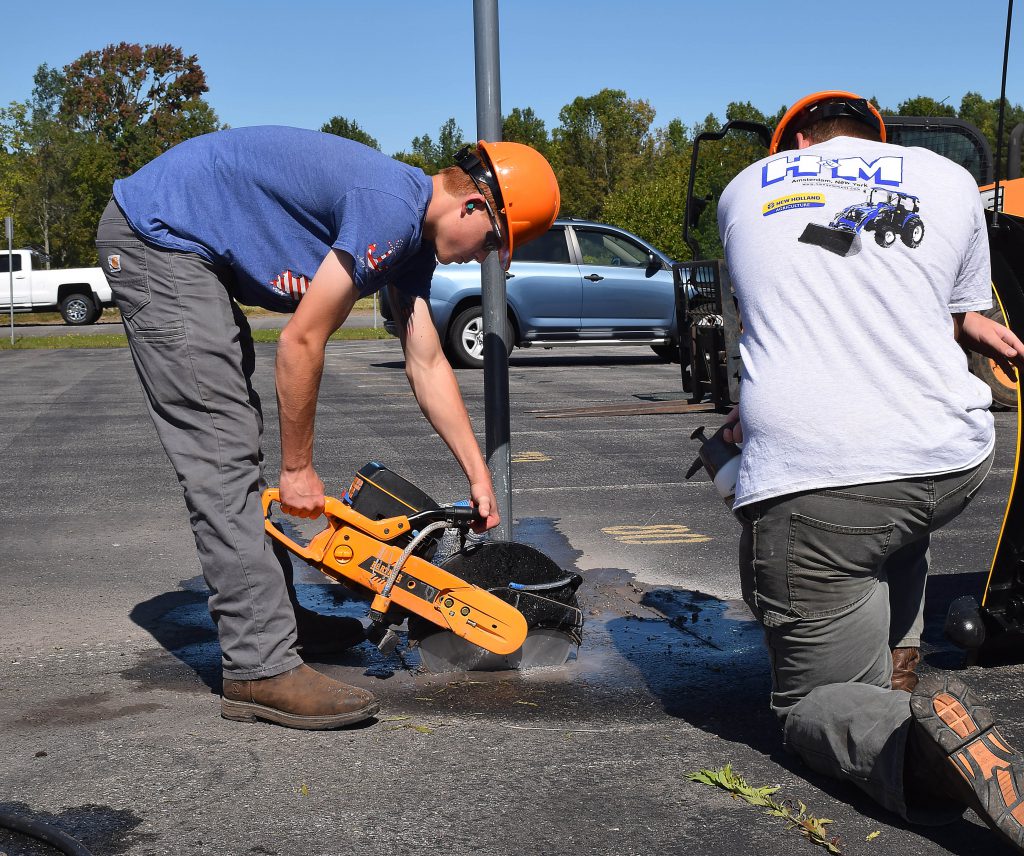 A student uses a demolition saw to cut blacktop around a metal fence post