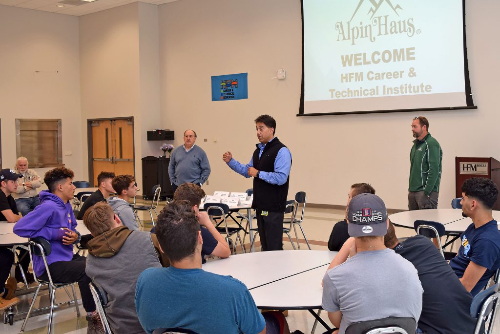 Three men stand in the front of a large room with Alpin Haus displayed on a large screen.