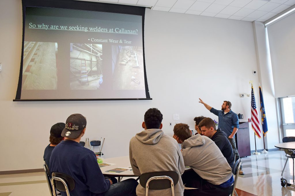 A speaker points to a Powerpoint presentation displayed on a screen as student sit at a table listening. 