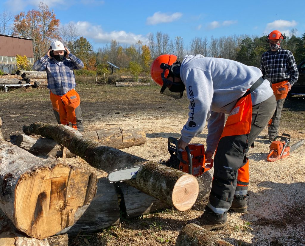 A student cuts a log with a chainsaw as an instructor and another student look on.