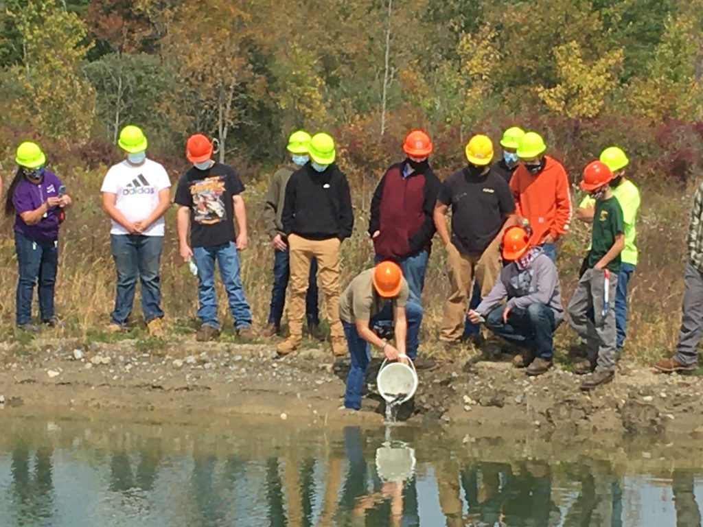 Students stand by a pond as an instructor add fish from a bucket