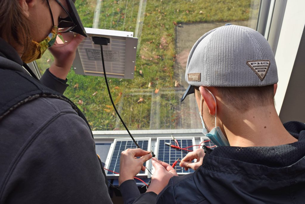 Two students stand next to a window in the sun and wire small solar panels.