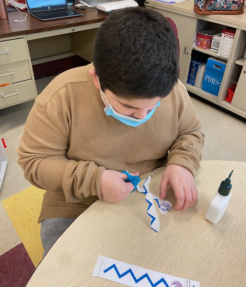A students uses scissors to cut paper with a glue bottle next to him.