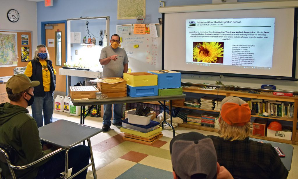 Jeff Fagan and Kathryn Gulick talk to a class showing a powerpoint with bee boxes in front of them