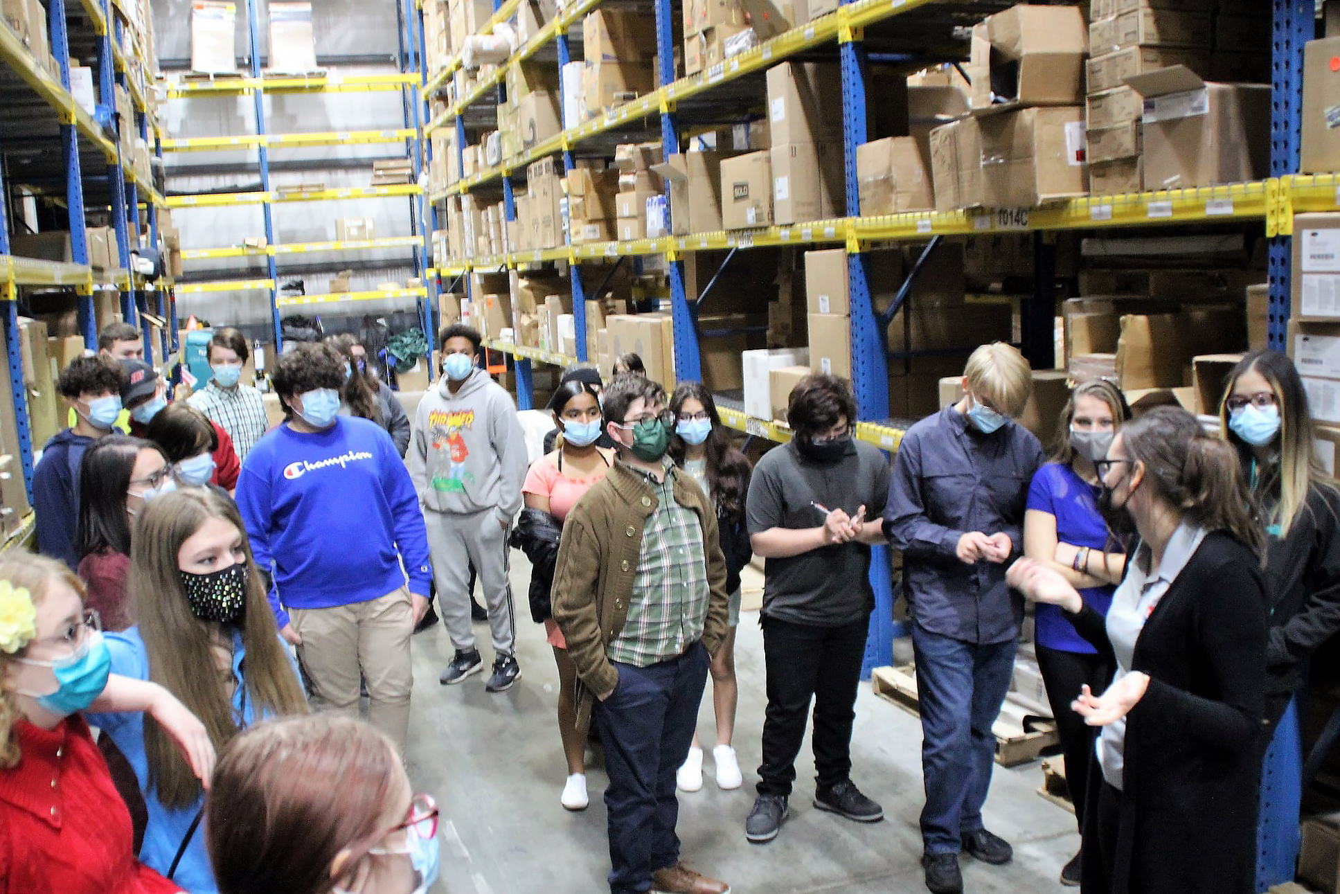 A group of students listens to a employee as they walk through the stacks of a warehouse