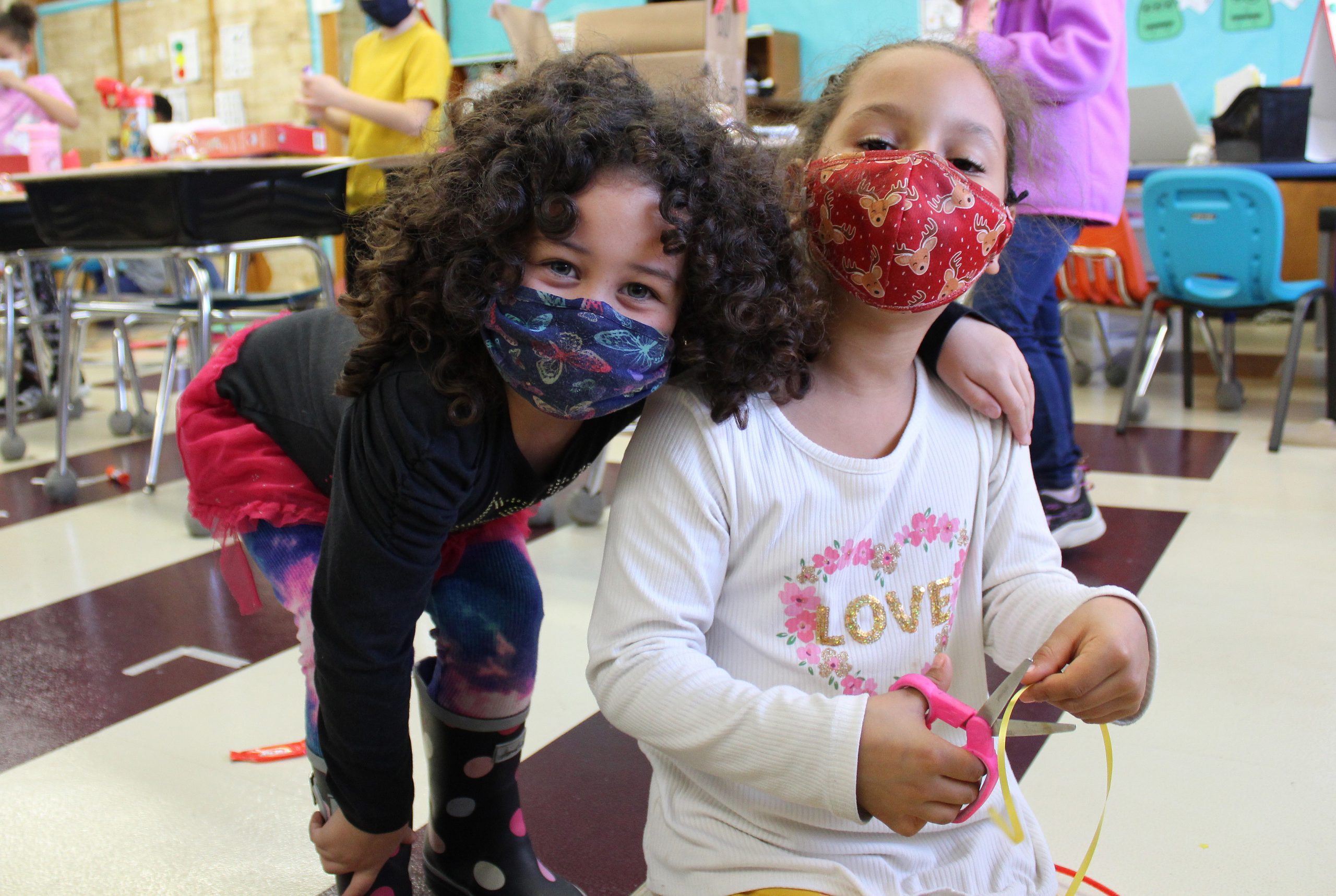 a student puts her arm around another student who is doing a craft project with scissors.