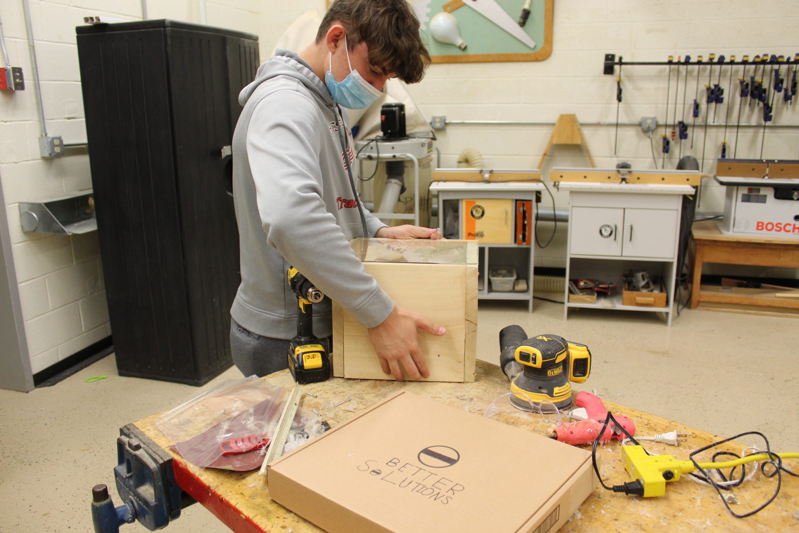 A student in a shop constructs a box with a drill and sander nearby
