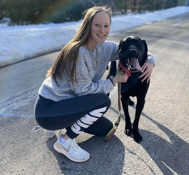 Kelsey kneels next to a black lab dog with it's tongue hanging outdoors