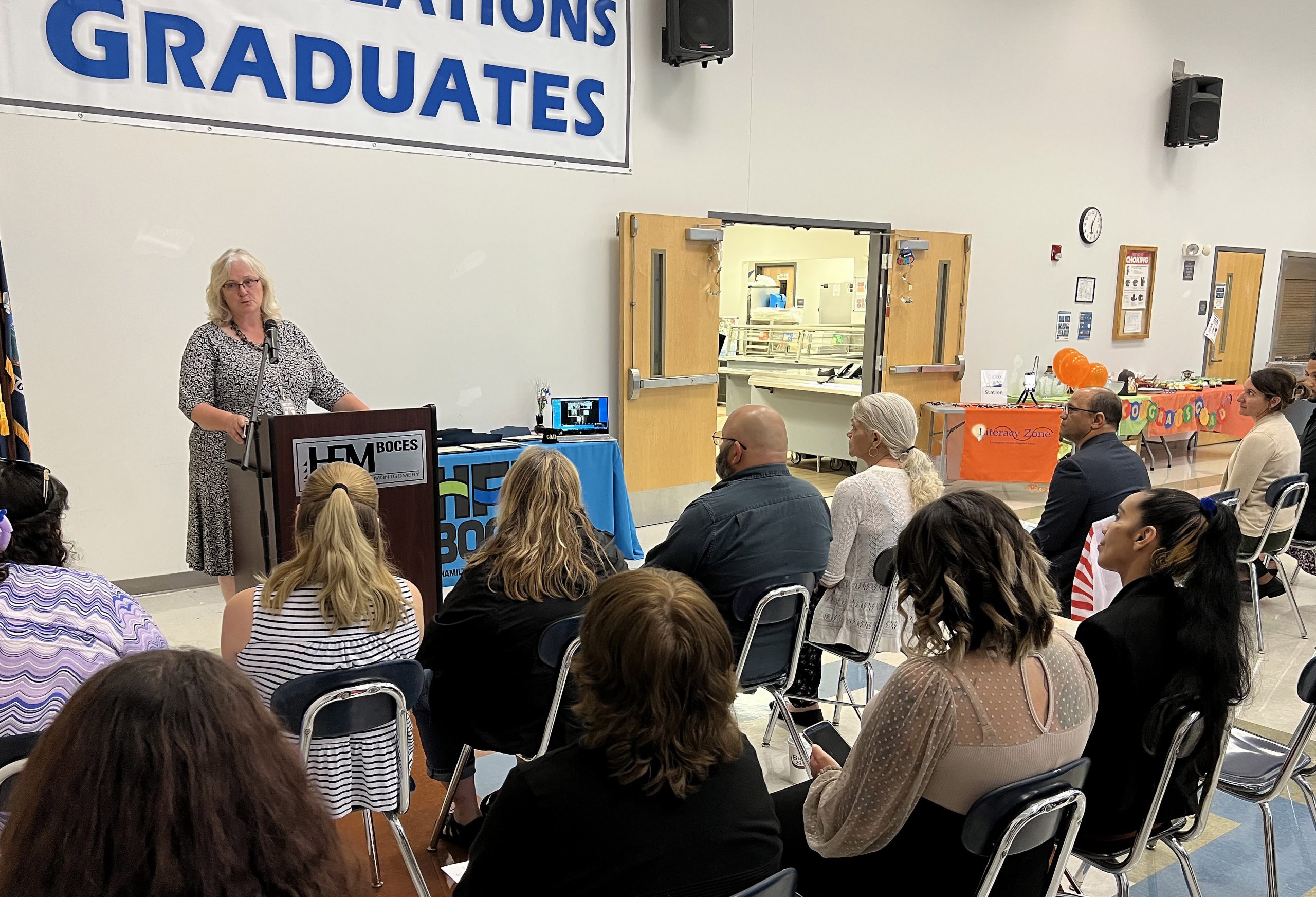 Adult Education Coordinator Laurie Bargstedt stands at a podium speaking to a crowd of adult graduates and family members