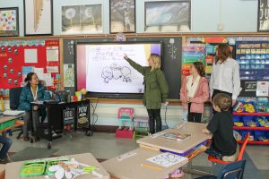 A PTECH student points to a Promethean board while discussing a book she wrote with her classmates in Chinese
