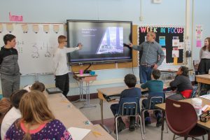 Two PTECH students point to Promethean board while educating elementary school students about a landmark in China