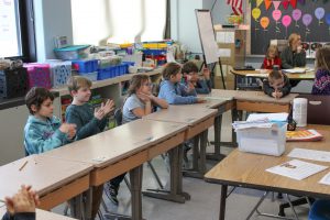 Group of elementary school students sitting at a long table listening to presentation being give in Chinese language by PTECH students