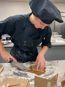 A student cuts gingerbread with a chef's knife to form a wall of her project.