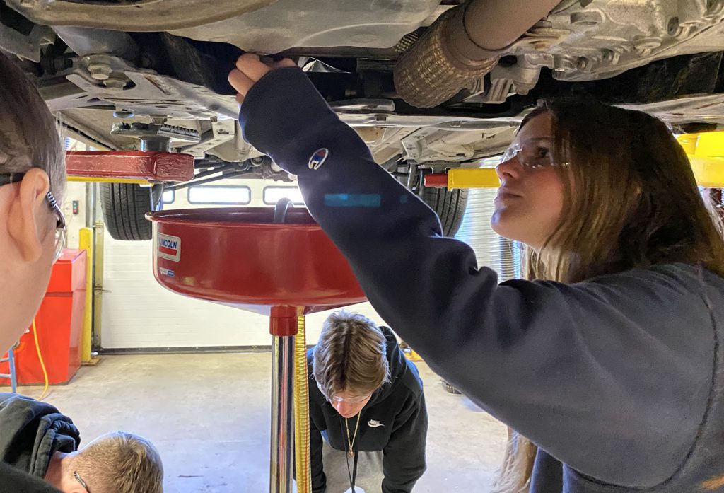 Several students stand under a car on a lift, while one student removes the oil plug.
