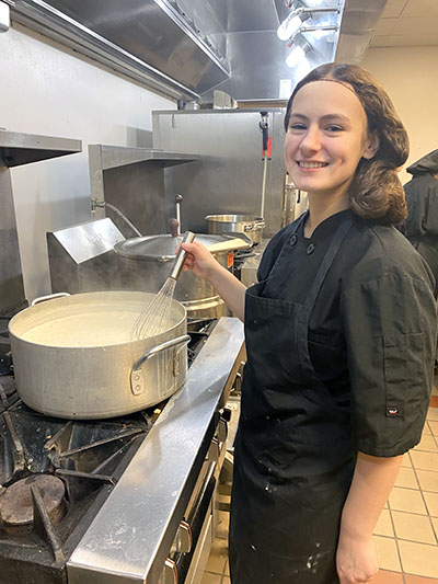A student stirs a big pot with a whisk and smiles at the camera.