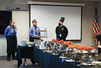 Two culinary students stand next to a buffet while their instructor looks on.