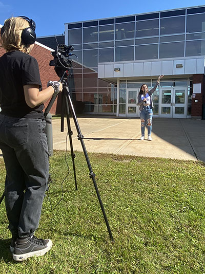 A student stands behind a video camera, filming another student who is holding a microphone. 