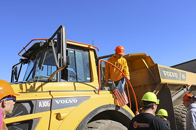 A student wearing a hard hat climbs down from the cab of a large dump truck.