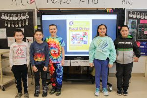 Students stand in a classroom while a promotional flier about the food drive is shown on a whiteboard behind them.