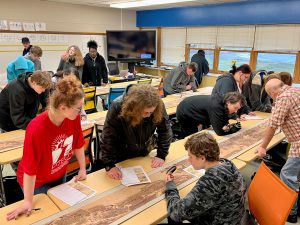 PTECH students are gathered around a desk in a classroom examining the ancient Chinese scroll titled Qingming Shanghe Tu