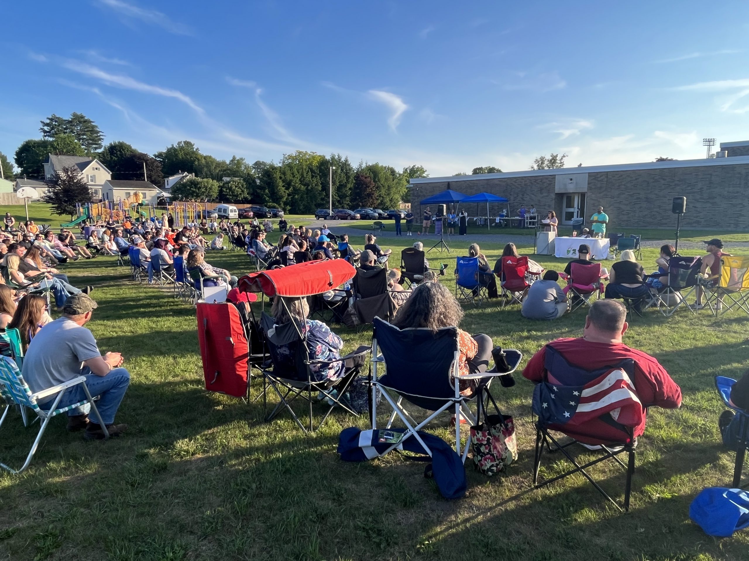 group of people in lawn chairs listen to a speaker at a podium