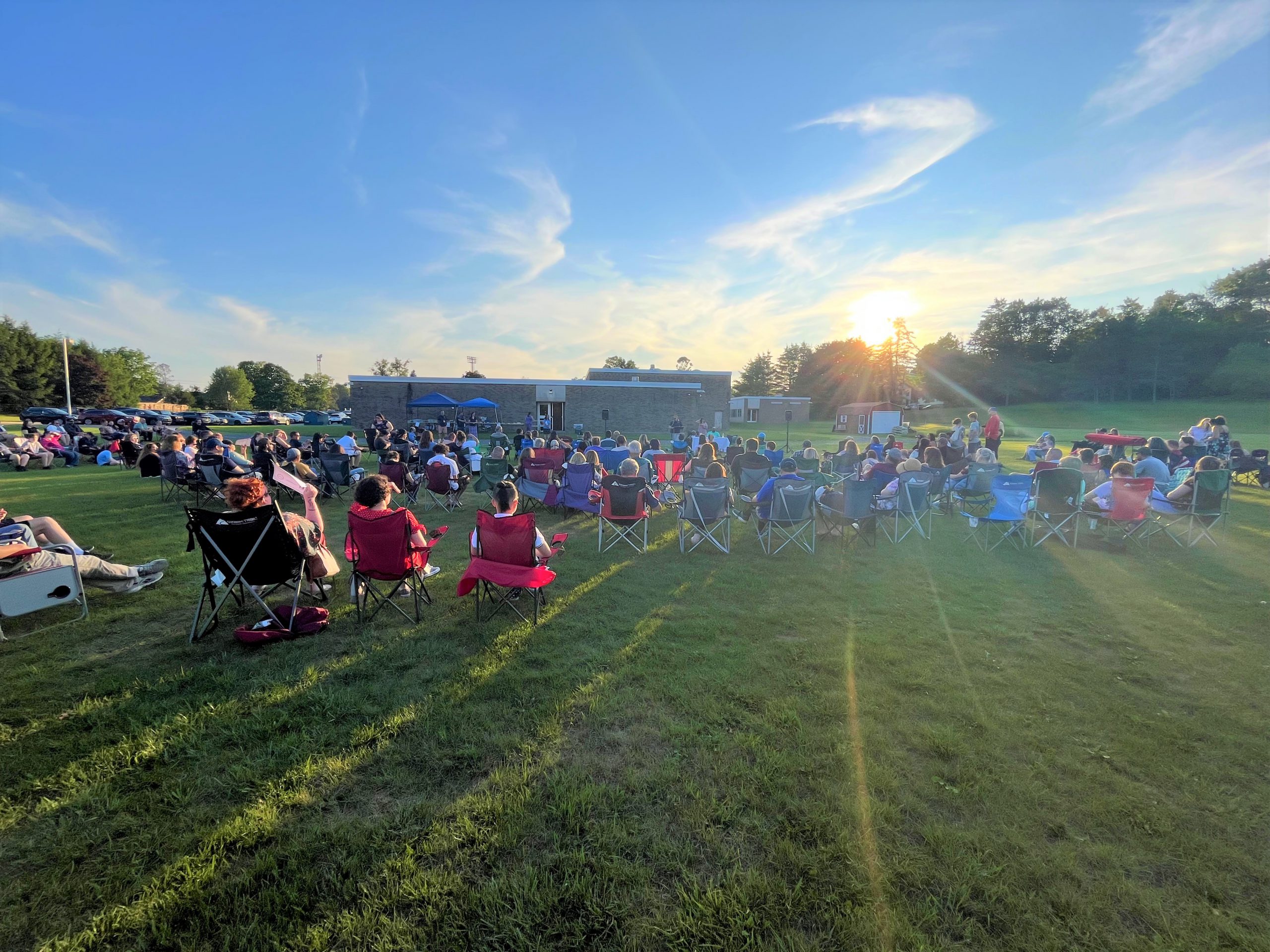 large group of people sit in lawn chairs and listen to a speaker with a sunset in the background