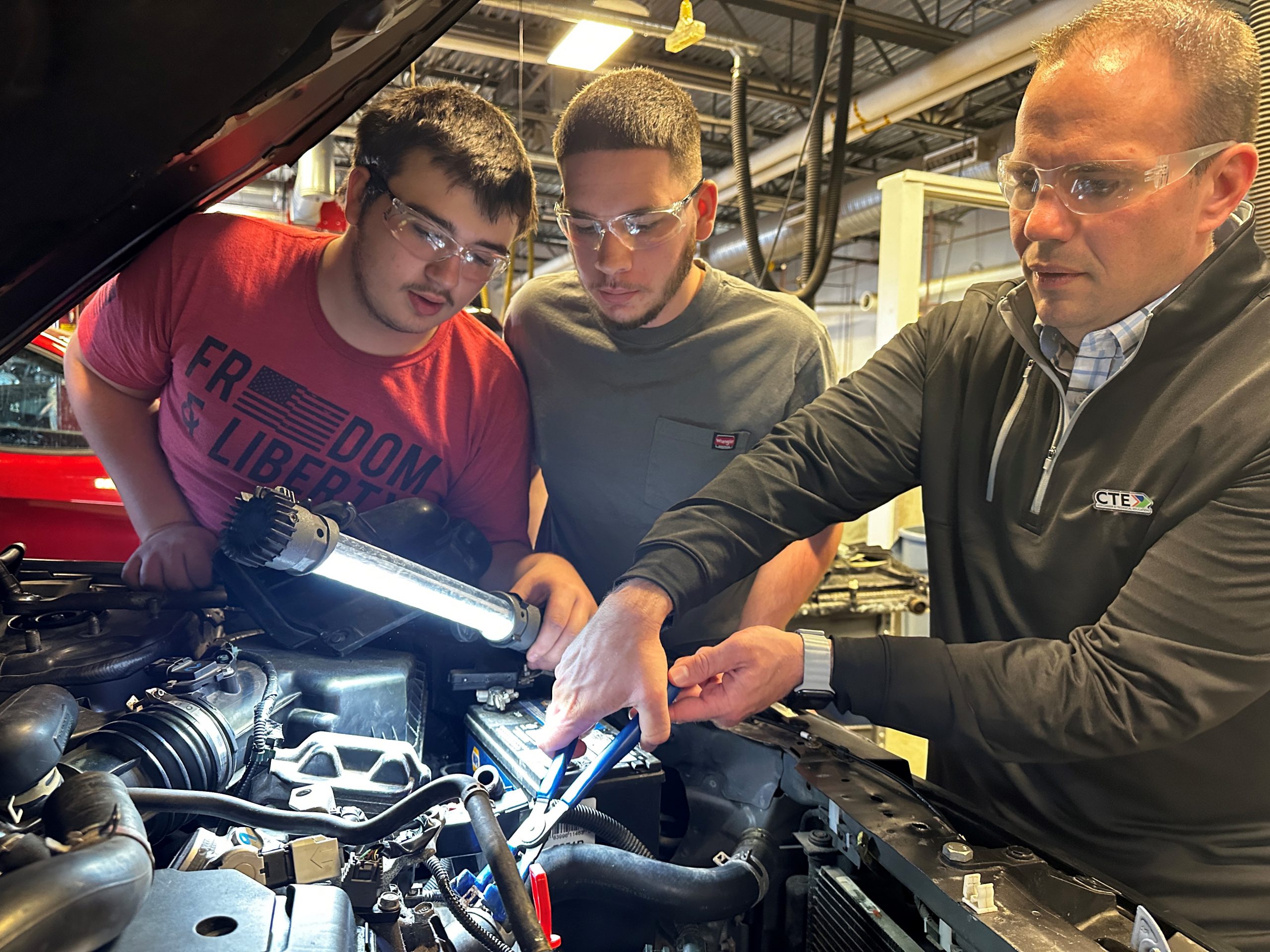 Mike Ott, far right, is pictured working under the hood of a vehicle in the CTE Automotive Technology program. He has a tool in his hand and is being guided by two students who are standing to his side.