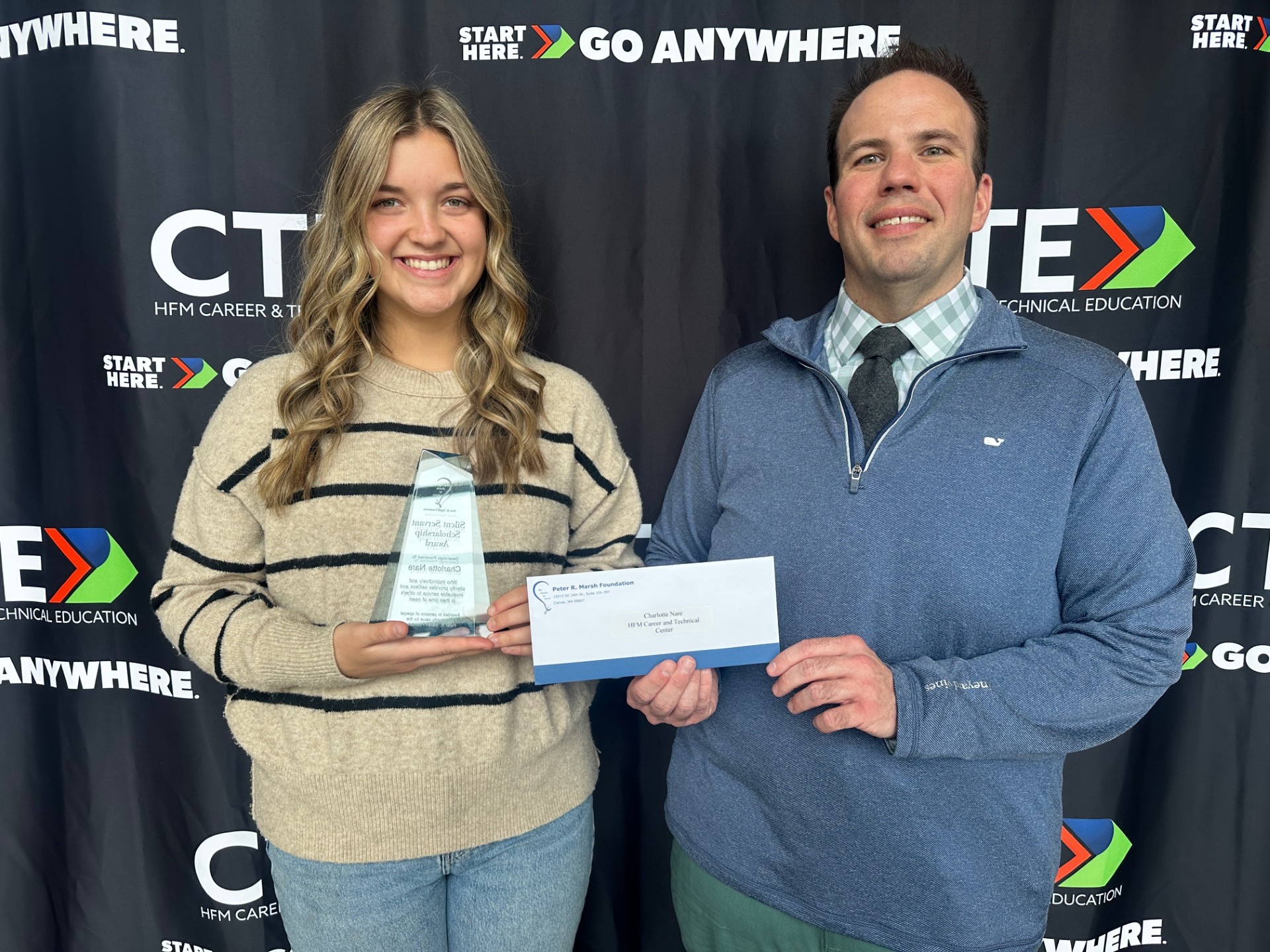 Charlotte Nare (left) and Michael Ott (right) pose for a picture in front of a black backdrop. Nare is holding the silent servant award and Ott is holding an envelope with a check inside that Nare received for being selected as one of two HFM CTE students to receive the Peter R. Marsh Foundation Silent Servant Scholarship Award.