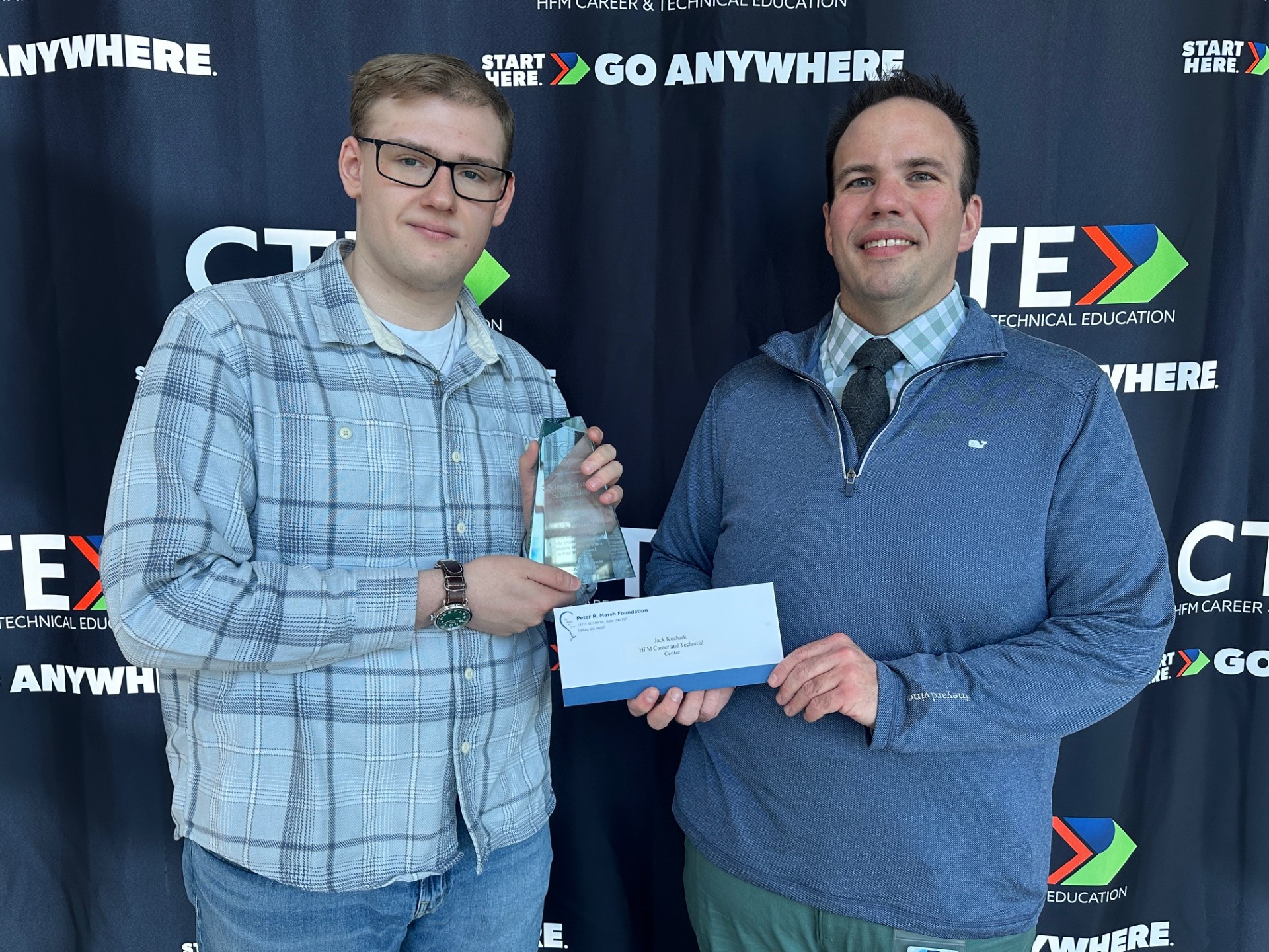 Jack Kuchark (left) and Michael Ott (right) pose for a picture in front of a black backdrop. Kuchark is holding the silent servant award and Ott is holding an envelope with a check inside that Kuchark received for being selected as one of two HFM CTE students to receive the Peter R. Marsh Foundation Silent Servant Scholarship Award.