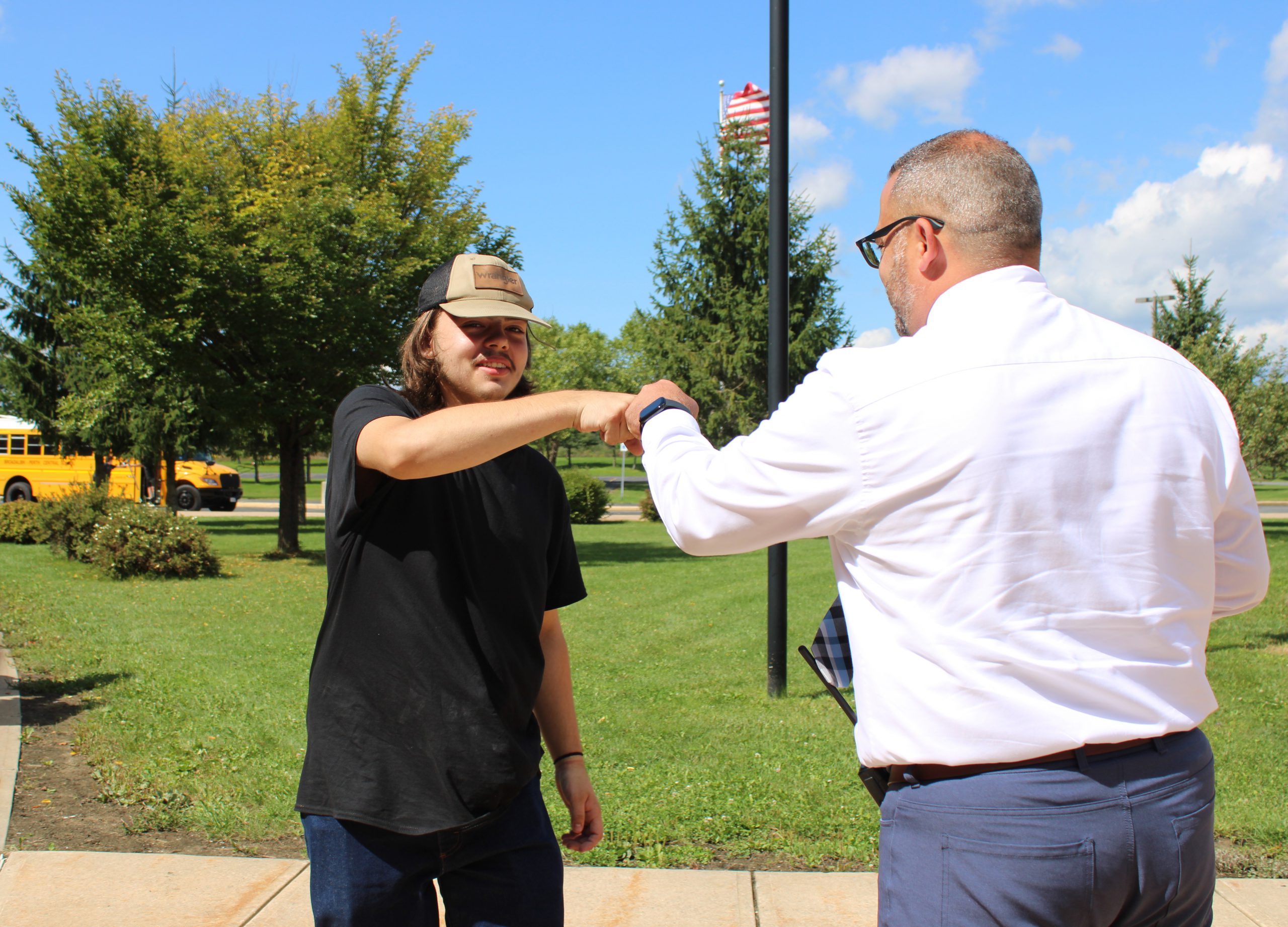 A student and an HFM CTE staff member greet each other with a fist bump outside the HFM Career and Technical Education Center. The student stands on a sidewalk near a grassy lawn and trees, while yellow school buses are visible in the background on a sunny day. An American flag can be seen flying behind them.